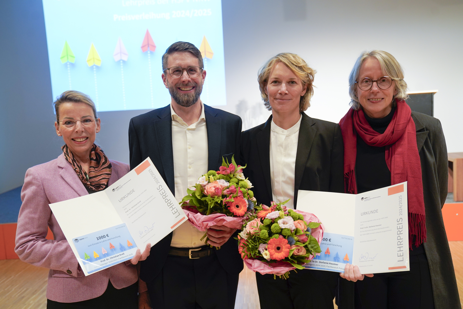 Iris Wiesner und Maria Flück stehen links und rechts am Rand und halten die offenen Urkunden hoch. Christian Krell und Stefanie Haumer stehen jeweils mit einem Blumenstrauß zwischen ihnen.