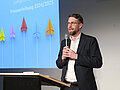 Lehrpreis 2025 Christian Krell am Rednerpult vor einer Beamerprojektion. In beiden Händen hält er ein Mikrofon.