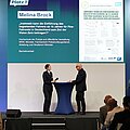 Melina Brock steht links auf einer Bühne an einem Stehpult. Gegenüber von ihr auf der rechten Seite steht der Moderator. Im Hintergrund wirft ein Beamer das Thema ihrer Thesis an die Wand.